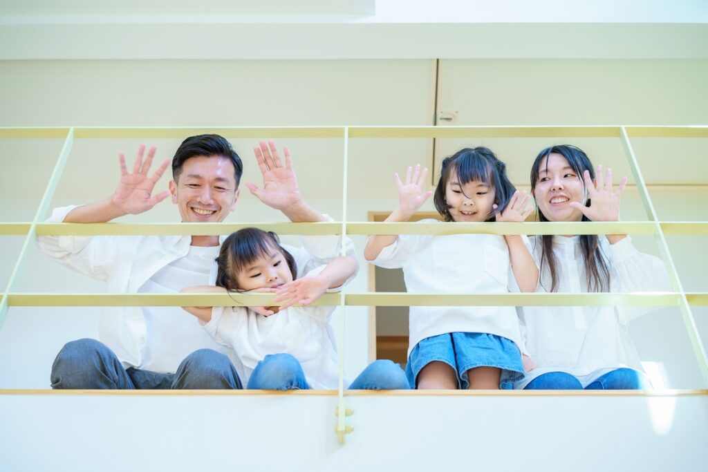 室内の手すり越しに並び、笑顔で手を振る家族の様子が写る明るい住空間