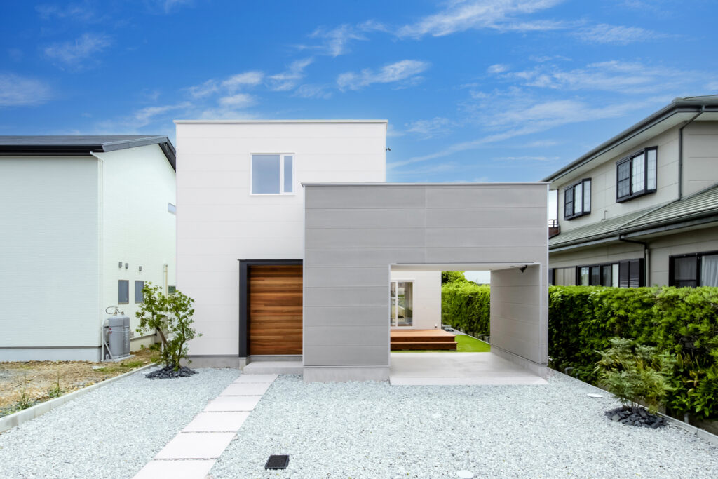 白とグレーの塗り壁を組み合わせたモダンなボックス型の外観。玄関周りに配された天然木のアクセントと、白砂利に敷石を並べたスタイリッシュなアプローチが調和する機能美。