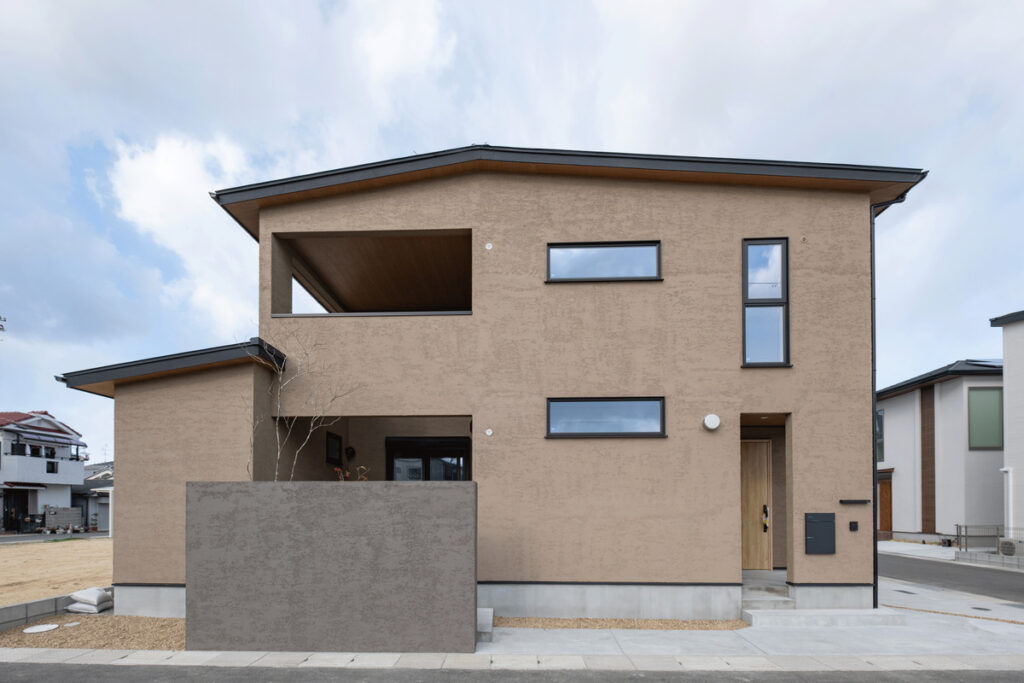 アースカラーの塗り壁と黒いサッシが調和する、シンプルモダンな外観デザイン。高い目隠し壁がプライバシーを守り、重厚感のある佇まいを演出する。