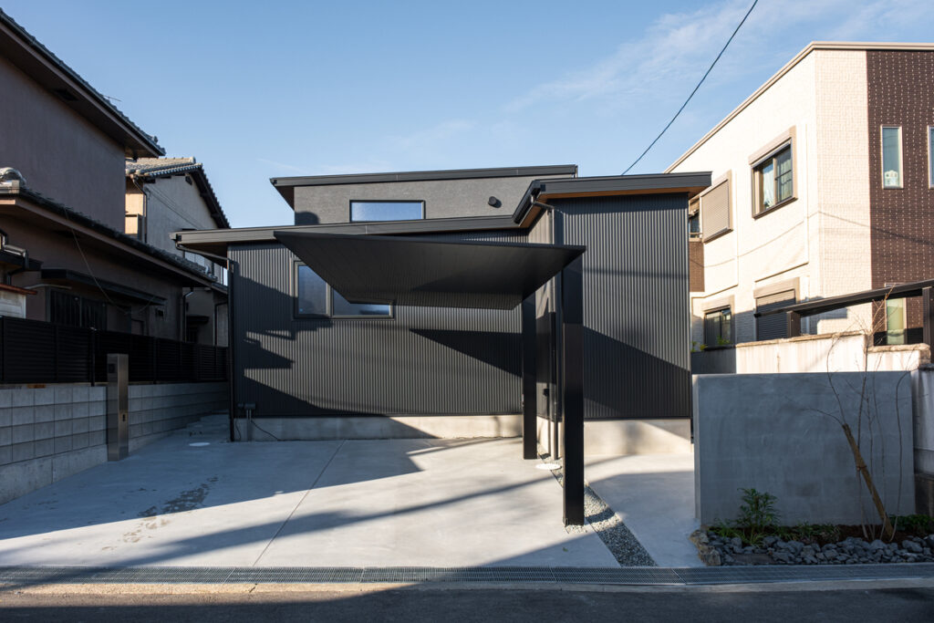 黒の縦平葺き外壁にフラットデザインのカーポートが美しく調和する、都会的でミニマルなスタイルの住宅外観。