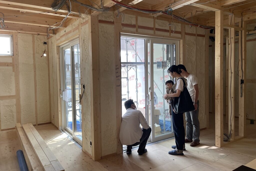 壁面に吹き付け断熱が施された建築現場で、大きな窓の施工状態を担当者と共に確認する施主家族。