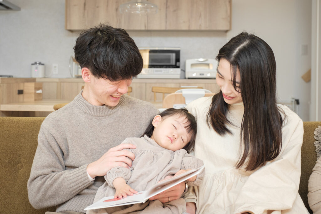 木の温もりを感じるキッチンを背景に、リビングのソファで眠る幼い子どもをパパとママが優しく見守る幸せなひと時。