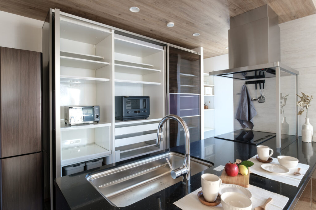 黒の石目調天板が映えるキッチン。背面の大型スライド収納には家電が機能的に収まり、天井の木目クロスが温かみを添えている。