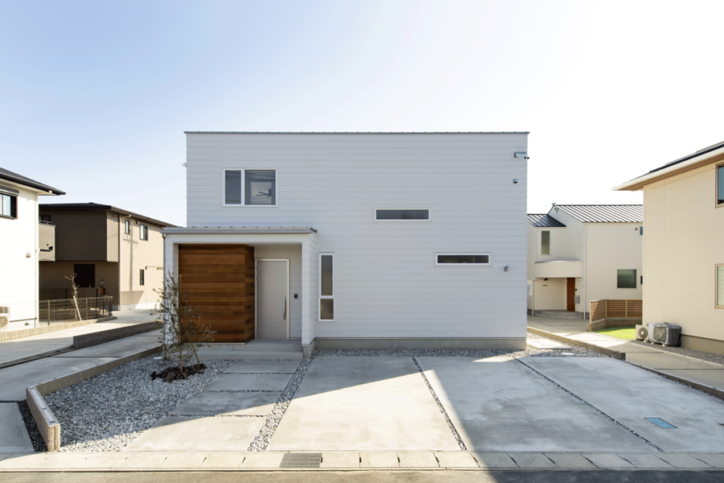 直線美が際立つグレーの塗り壁外観。玄関ポーチの木目調アクセントが温かみを添え、コンクリート舗装の駐車場と白砂利で整えられた、機能的で清潔感のあるオープン外構。