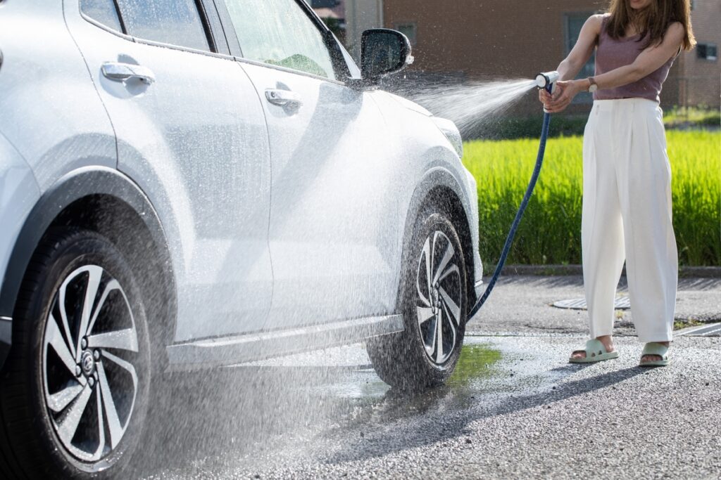 晴天の下、ホースから勢いよく水を出し、愛車の汚れを洗い流してメンテナンスを楽しむ休日の一コマ。