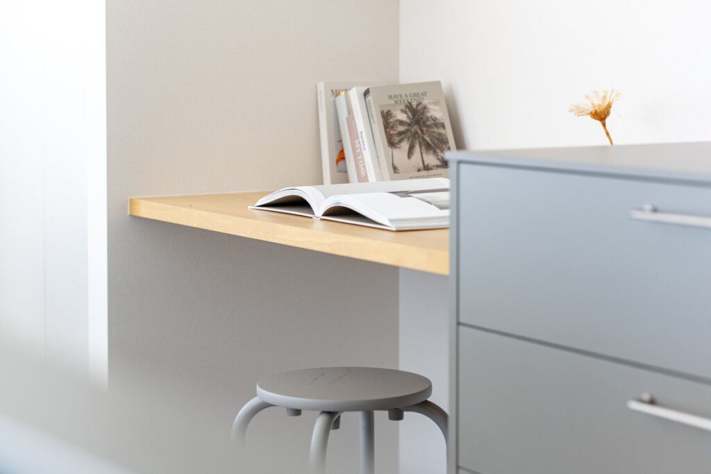 部屋のコーナーに設けられた造作の木製デスクと、グレーの丸椅子やチェストが調和する、シンプルで洗練されたワークスペース。