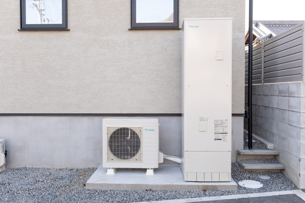 住宅の外壁横に設置されたエコキュートのヒートポンプユニットと貯湯タンク。コンクリート架台に据え付けられたオール電化の設備。