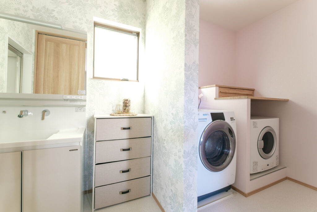 ボタニカル柄の壁紙が映える洗面室。奥にはドラム式洗濯機と乾燥機をカウンター下に配置した、家事効率の良いランドリー。