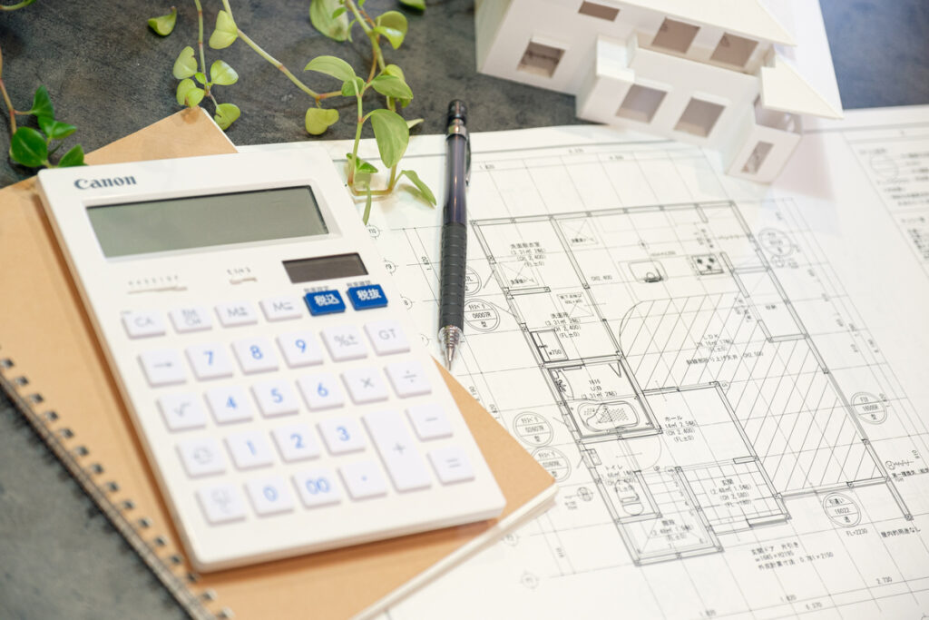 住宅設計図面の上に置かれた電卓、ペン、白い建築模型。具体的な家づくりの計画や資金シミュレーションを行う様子。
