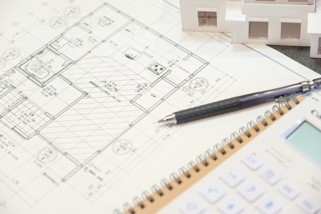 注文住宅の間取り図、住宅模型、電卓、筆記用具が机に並んだ、夢のマイホームに向けた詳細なプランニングをイメージさせる一枚。