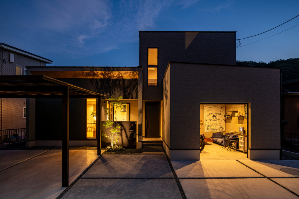 夜の静寂に浮かび上がる、ライトアップされた美しい外観。右側のインナーガレージは趣味を楽しめる部屋として活用され、温かなオレンジ色の光がモダンな建物を彩る幻想的な風景。