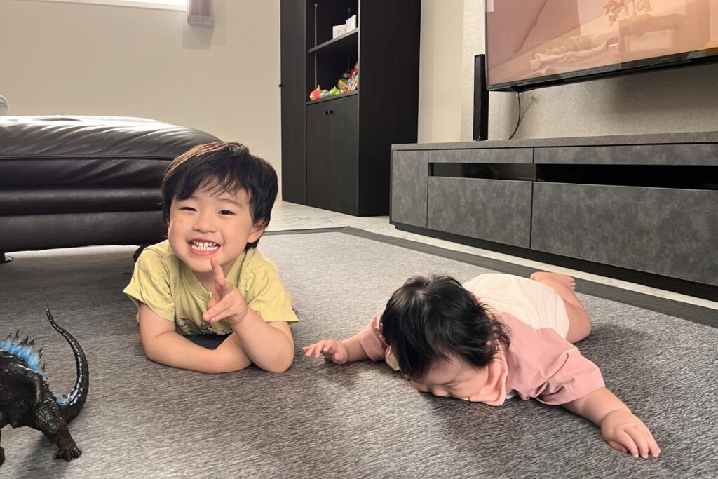 リビングのカーペットの上で、おもちゃに囲まれながらリラックスして遊ぶ笑顔の男の子とハイハイをする赤ちゃんの日常風景。