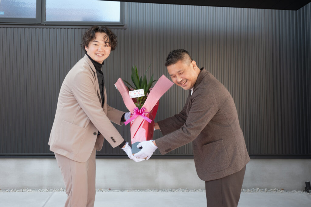 住宅の完成を祝い、笑顔で握手を交わしながらお祝いの品を贈呈する、信頼関係の深まりを感じさせる引き渡し式のワンシーン。