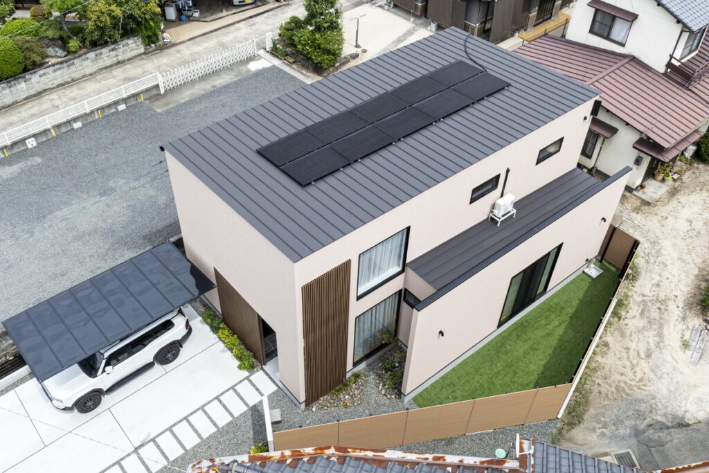 太陽光パネルを搭載したモダンな外観。ウッドルーバー、人工芝の庭、カーポートを備えた都市型住宅を上空から俯瞰した様子。