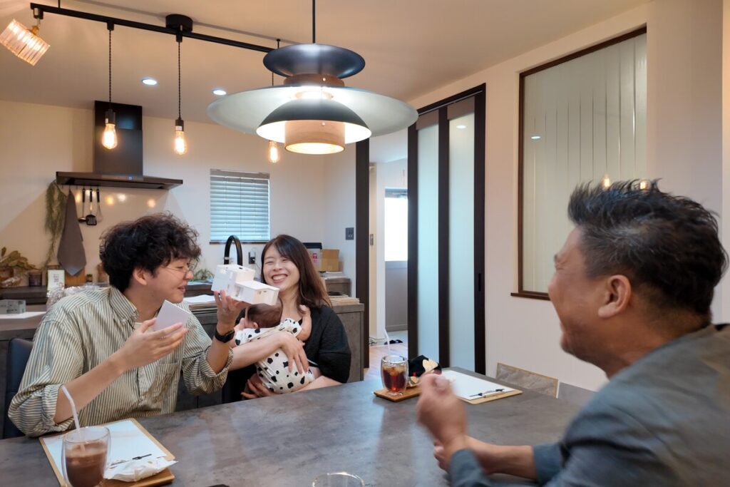 住宅模型を手に笑顔で打ち合わせを行う家族と工務店スタッフ。ダイニングのペンダントライトの下で、新しい住まいへの期待を膨らませながら相談を進める和やかな相談風景。