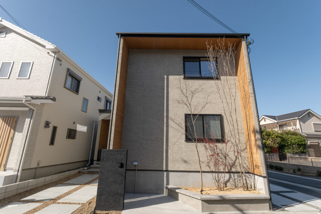 ベージュの塗り壁に木目調のアクセント壁が映える、洗練されたモダンな外観デザイン。スクエアなフォルムと繊細な植栽が調和する佇まい。